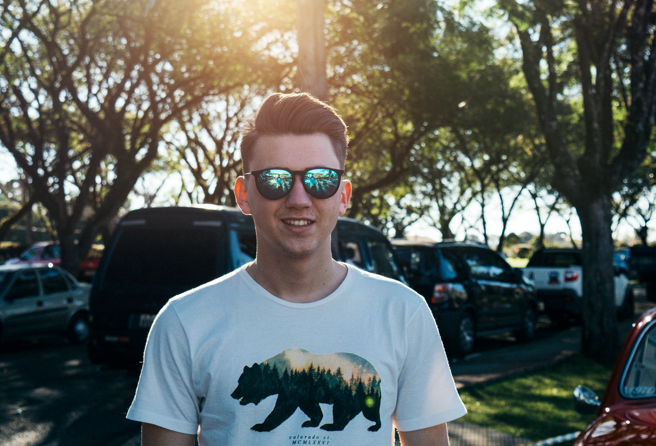 services-06 A young man in a graphic t-shirt and sunglasses smiles outdoors with blur background.